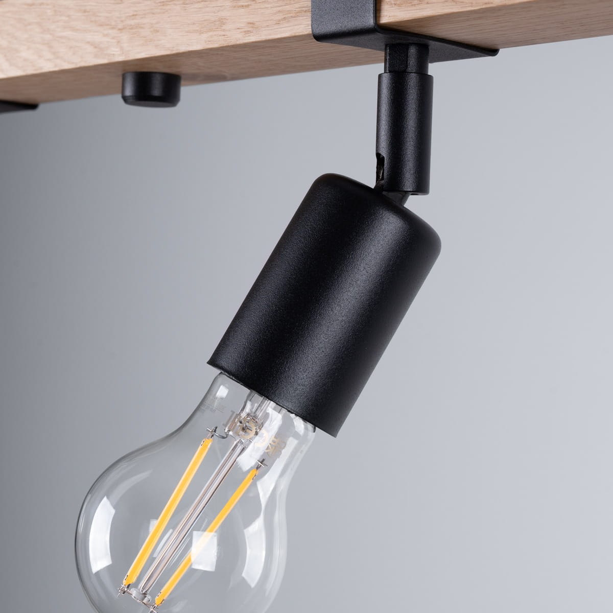 Czarna lampa sufitowa Zuca z miejscem na 4 żarówki E27, zamontowana pod drewnianym stropem. Widoczny detal uchwytu w kształcie cylindrycznym, z przezroczystą żarówką, podkreślającą nowoczesny styl oświetlenia. Tło w odcieniach szarości.