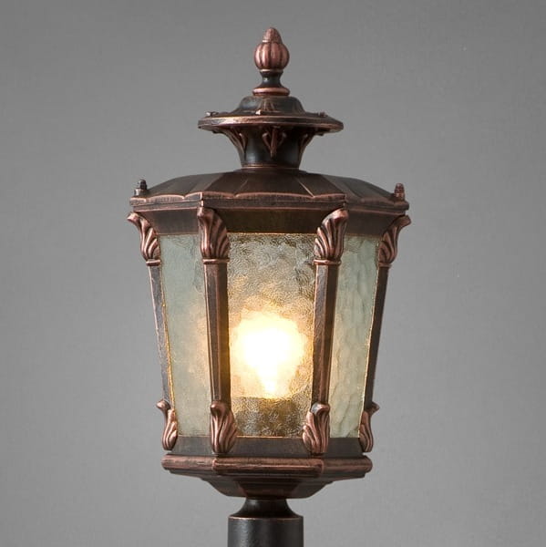 Lampa stojąca ogrodowa Amur 1-punktowa, w kształcie latarni. Metalowa obudowa w kolorze mosiężnym z dekoracyjnymi detalami, szklane ścianki w stylu witrażowym. Źródło światła widoczne w środku, całość na ciemnym tle.
