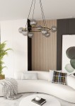 Lampa wisząca Azura 8xE14 w kolorze czarnym i grafitowym. Posiada okrągłą konstrukcję z ośmioma szklanymi kulami w odcieniu szaro-srebrnym, zawieszona na cienkich czarnych linkach. Umieszczona w nowoczesnym wnętrzu z jasną sofą i roślinnością.