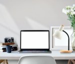 Lampka biurkowa Senza w kolorze białym, z metalowym kloszem i drewnianą podstawą. Ustawiona obok laptopa na biurku, w otoczeniu książek, kubka i kwiatu w wazonie. Kąt ujęcia z boku, ukazujący detale lampki oraz aranżację biura.