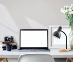 Lampka biurkowa Senza w kolorze czarnym z podstawą z naturalnego drewna. Posiada elastyczną szyjkę i oprawę na żarówkę E27. Ustawiona na biurku obok laptopa, filiżanki i książek, w tle z dekoracjami.