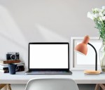 Lampka biurkowa Senza 1xE27 w kolorze pomarańczowym, z metalowym ramieniem i drewnianą podstawą, ustawiona na biurku obok laptopa, książek i filiżanki, w jasnym wnętrzu z kwiatami w wazonie.