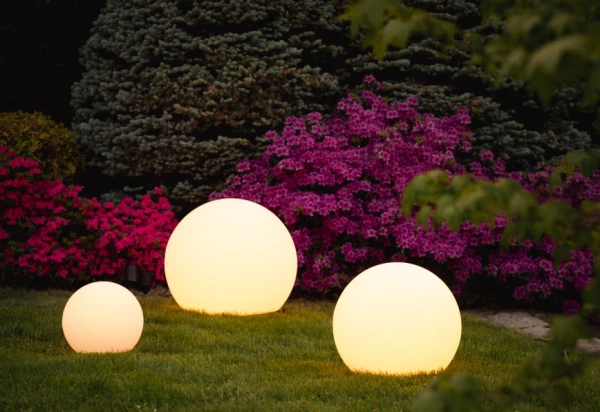 Lampa ogrodowa Cumulus L w kształcie kul, biała, z źródłem światła E27. Umieszczona na trawie, obok kolorowych kwiatów. W zestawie trzy kule o różnych rozmiarach, tworzące dekoracyjną aranżację świetlną w ogrodzie.