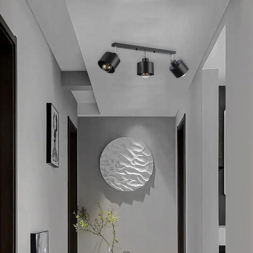 Lampa sufitowa Elza w kolorze czarnym, montowana na suficie, z trzema reflektorami E27 w różnych pozycjach. Prostokątna podstawa, metalowa, z widocznymi detalami. Otoczenie w odcieniach szarości, minimalistyczny styl.