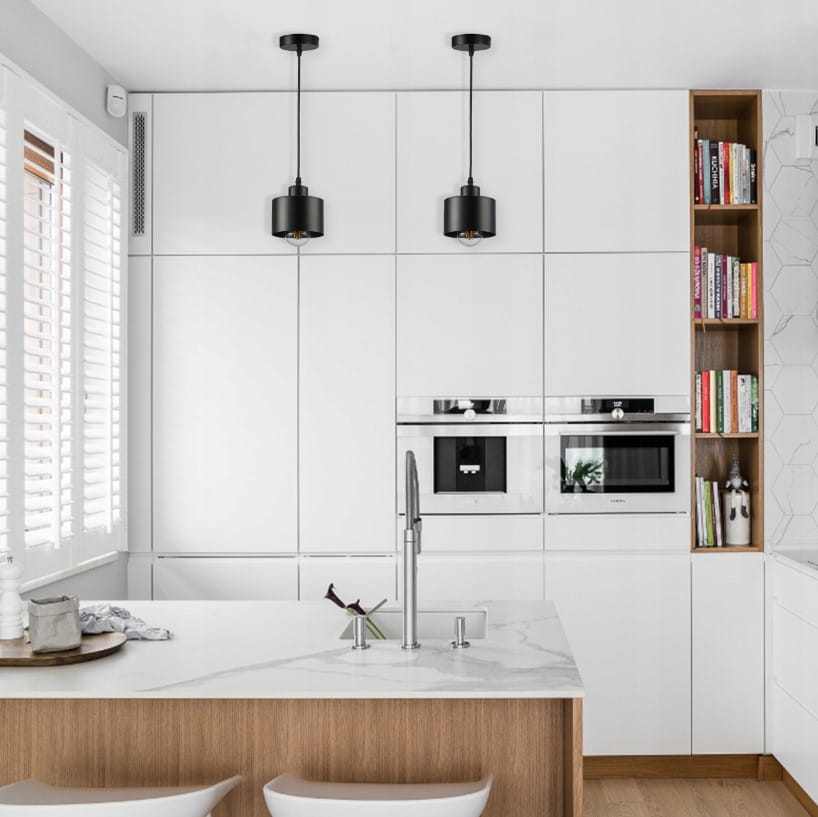 Czarna lampa wisząca Elza z pojedynczym gniazdem E27, zawieszona na długim kablu, umiejscowiona w jasnej kuchni z białymi szafkami i drewnianym blatem. W tle widoczny fragment kuchennego wyposażenia oraz półka z książkami.