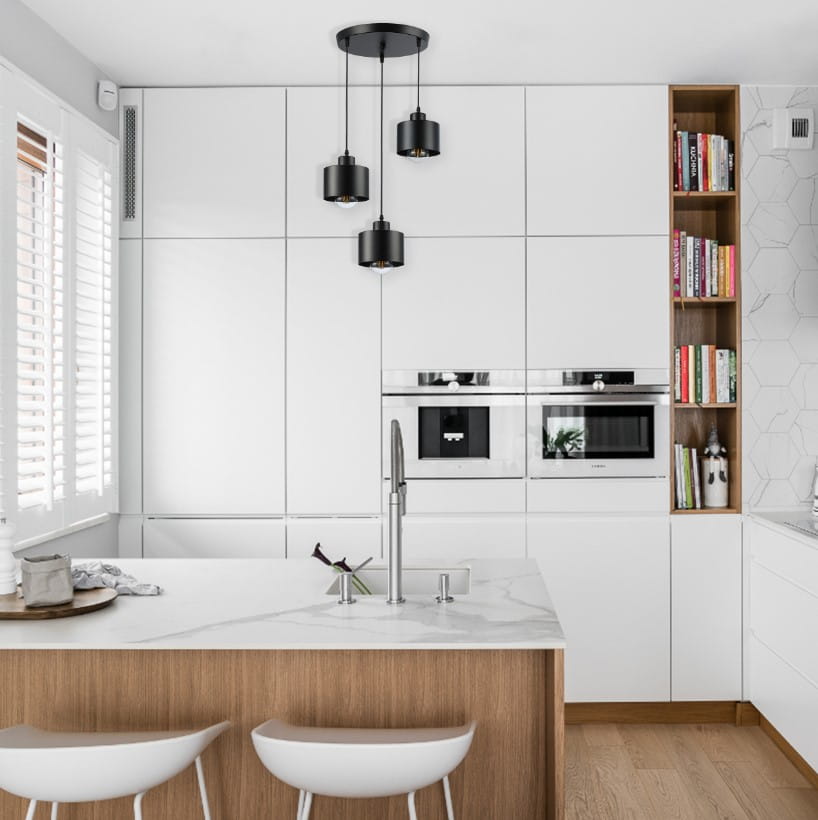 Czarna lampa wisząca Elza, okrągła, z trzema źródłami światła E27, zawieszona w kuchni nad wyspą. Lampa ma minimalistyczny design, z metalowymi kloszami w kształcie cylindrów, umieszczona na czarnej podsufitce. Tło stanowi jasna zabudowa kuchenna.