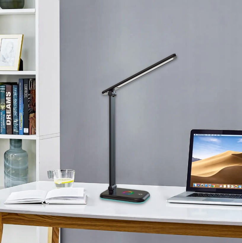 Czarna lampka biurkowa Alma LED 12W/CCT + RGB, z regulowanym ramieniem, umieszczona na biurku obok laptopa. Na podstawie znajduje się kolorowy panel dotykowy. W tle widoczna półka z książkami i szklanka z wodą.