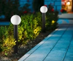 Lampa stojąca ogrodowa Feltre H80, czarna, z okrągłym kloszem w kolorze białym, umieszczona na podłużnym słupku. Widoczna wzdłuż ścieżki, otoczona zielonymi krzewami, tworzy oświetlenie w ogrodzie.