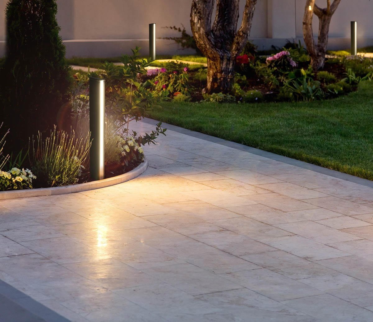 Lampa stojąca ogrodowa Grado 3K w kolorze czarnym, wysokość 70 cm, z jedną diodą LED 12W, 3000K, umieszczona na ścieżce w ogrodzie, otoczona roślinnością i kwiatami, oświetlająca teren w dolnej części, z prostym, cylindrycznym kształtem.