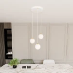 Lampa wisząca Royal 3-punktowa w kolorze białym i złotym, z trzema okrągłymi kloszami zawieszonymi na cienkich linkach. Ujęcie z góry, w nowoczesnym wnętrzu, na tle jasnych ścian i marmurowego stołu, z rośliną w doniczce.