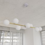 Lampa wisząca Gravity 8-punktowa z białymi, opalowymi kloszami na złotej listwie. Ujęcie z góry, z widocznymi sześcioma kulistymi kloszami w równych odstępach. Lampa zawieszona na białych przewodach, montowana do sufitu, na jasnym tle z zasłonami.