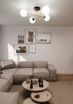 Lampa sufitowa 4-punktowa Magnum w kolorze czarnym, złotym i białym. Ułożona centralnie, z czterema białymi kulistymi kloszami na złotych ramionach. Tło z jasnymi ścianami, w aranżacji salonu z kanapą i dekoracjami na stoliku.