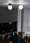 Lampa sufitowa Sopra, 2-punktowa, w kolorze czarnym z opalowymi kloszami. Ujęcie z góry, zamontowana na suficie, z widocznymi okrągłymi żarówkami. Tło w odcieniach szarości, z czarnym blatem i brązowymi krzesłami.