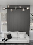 Lampa wisząca Pandora 8-punktowa w kolorze czarnym i grafitowym, z owalnymi, przezroczystymi kloszami, zawieszona nad białą sofą. Widok z boku, na tle szarej ściany. Klosze ukazują źródła światła, tworząc nowoczesny akcent w aranżacji.