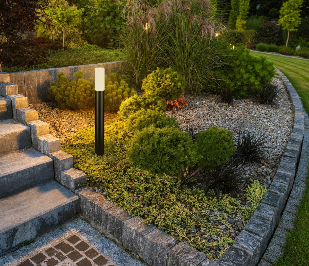 Lampa stojąca ogrodowa Muggia H80 w kolorze czarnym, z oprawą na jedną żarówkę E27. Ustawiona na tle zielonych krzewów i dekoracyjnych kamieni, oświetla ścieżkę w ogrodzie, tworząc przytulną atmosferę.