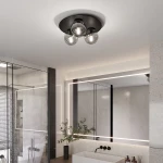 Trzypunktowa lampa sufitowa w kolorze czarnym i grafitowym, zamontowana na gładkim, jasnym suficie. Posiada trzy przezroczyste klosze, które eksponują źródła światła. Produkt znajduje się w nowoczesnej łazience, w otoczeniu lustra i rośliny w doniczce.