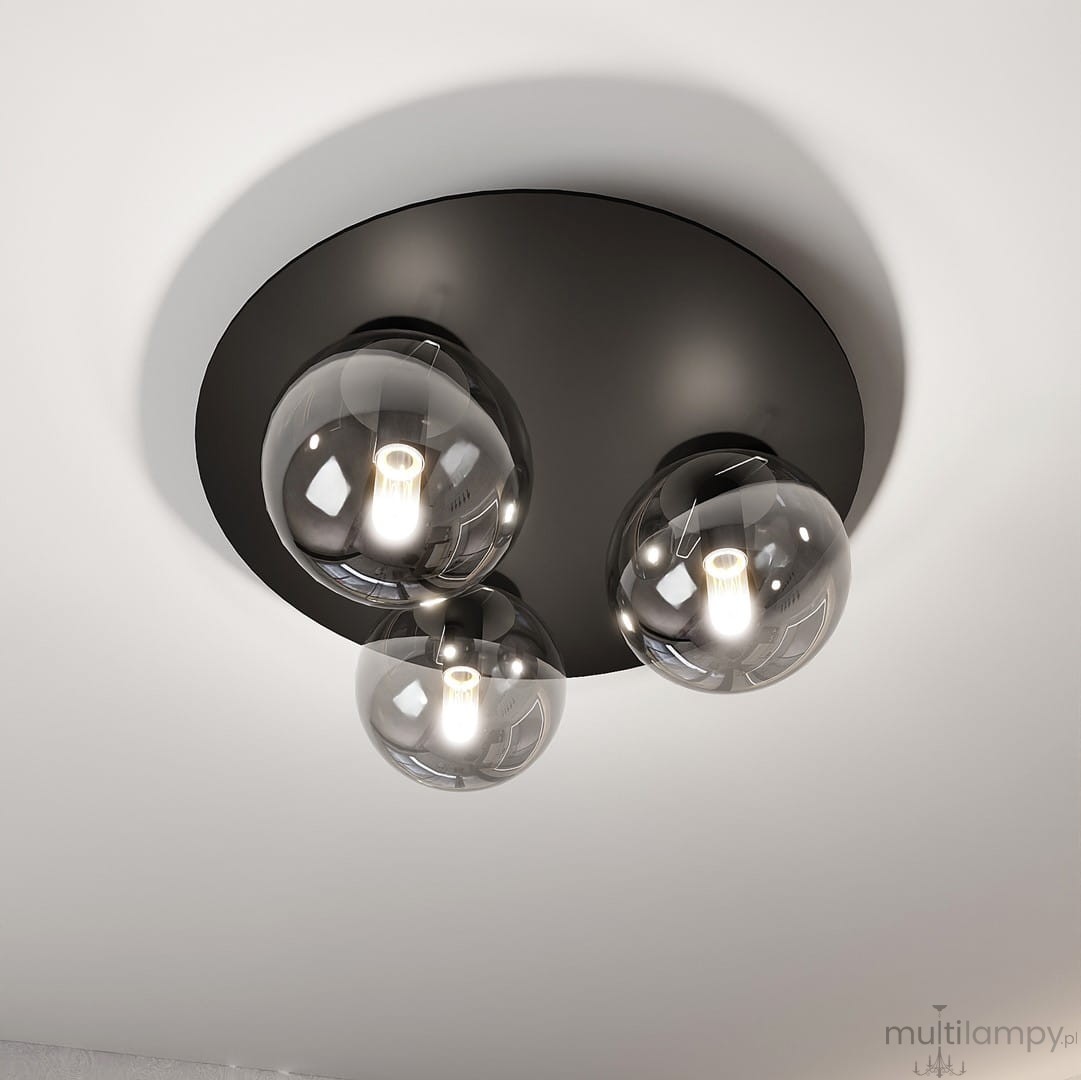 Trzypunkowa lampa sufitowa w kolorze czarnym z grafitowym stelażem. Okulary lamp wykonane z przezroczystego szkła, z widocznymi żarówkami w środku. Produkt zamontowany na okrągłej, matowej podstawie. Ujęcie z dołu, ukazujące całość lampy.