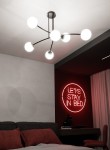 Czarna lampa wisząca Halldor 6-punktowa z okrągłymi, białymi kloszami, umieszczona na suficie. Klosze są rozmieszczone na elastycznych ramionach, tworząc nowoczesny design. W tle widoczny neonowy napis na czerwonym tle i łóżko z poduszkami.