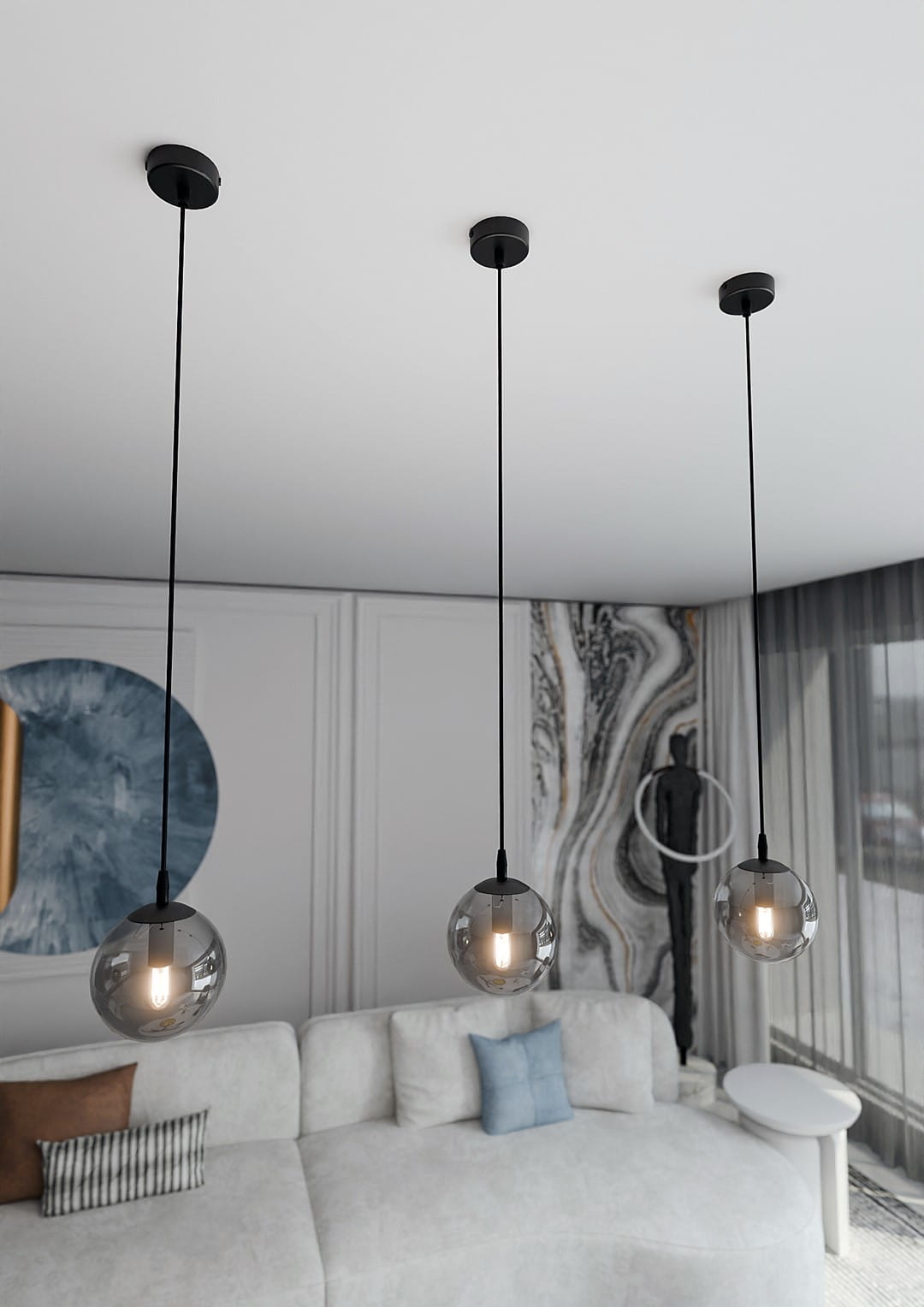 Grafitowa lampa wisząca Cosmo 1-punktowa z przezroczystą, okrągłą kloszem, zawieszona na cienkim, czarnym przewodzie. Ujęcie z góry, pokazujące lampę nad jasną sofą we wnętrzu. W tle minimalistyczne dekoracje, w tym abstrakcyjny obraz i rzeźba.