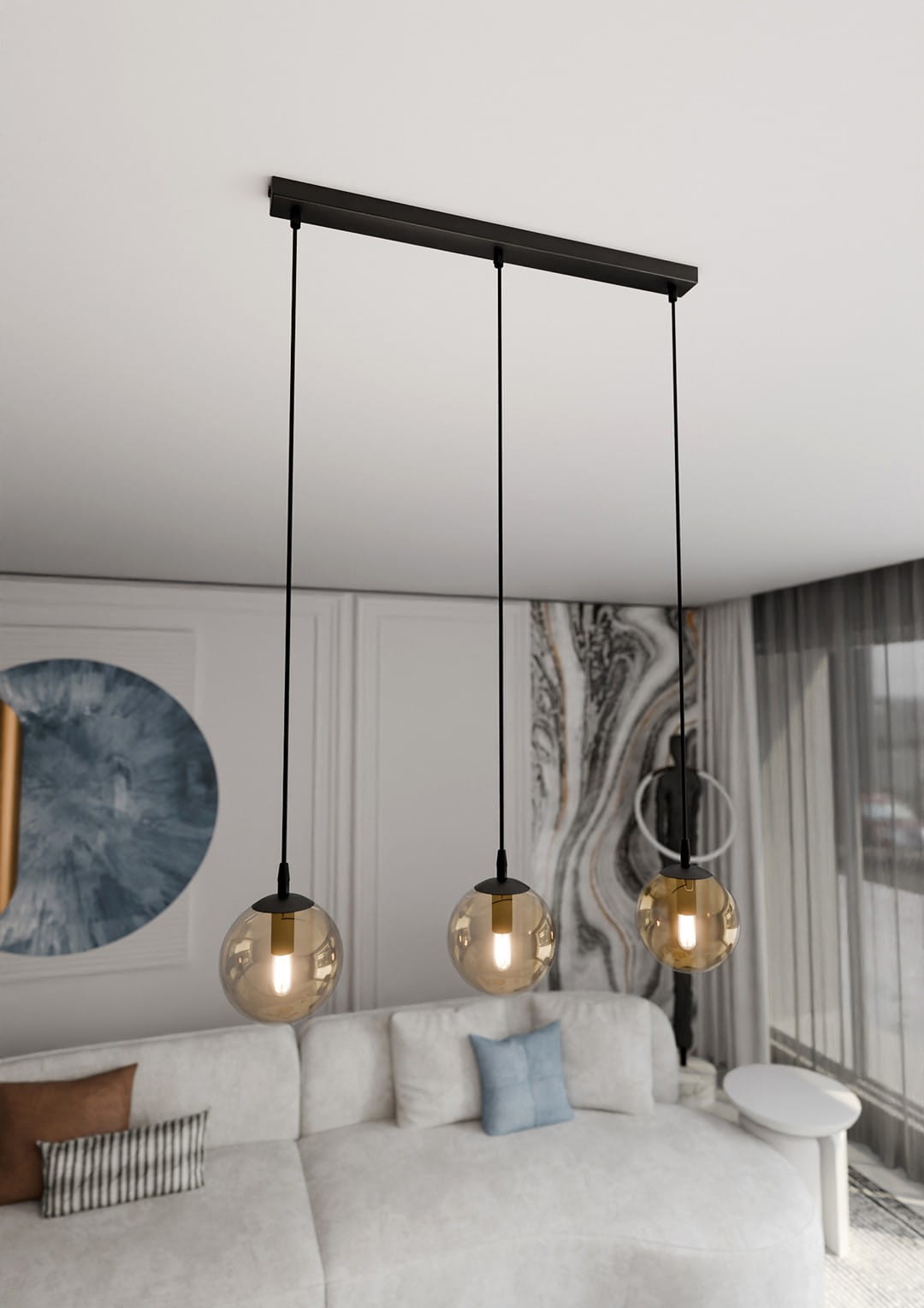 Lampa wisząca Cosmo 3-punktowa w kolorze miodowym, z trzema okrągłymi kloszami z przezroczystego szkła, zawieszona na czarnych linkach. Ujęcie z góry, z widocznym tłem w jasnych odcieniach, z kanapą i dekoracjami w salonie.