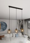 Lampa wisząca Cosmo 3-punktowa w kolorze miodowym, z trzema okrągłymi kloszami z przezroczystego szkła, zawieszona na czarnych linkach. Ujęcie z góry, z widocznym tłem w jasnych odcieniach, z kanapą i dekoracjami w salonie.