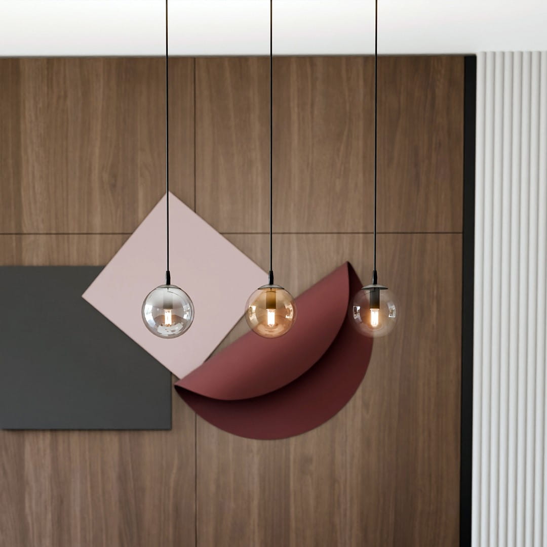 Lampa wisząca Cosmo 3-punktowa w multikolorze, z trzema okrągłymi kloszami w różnych kolorach (przezroczysty, brązowy, ciemny) zawieszona na czarnych linkach, w nowoczesnym wnętrzu z geometrycznymi elementami dekoracyjnymi na tle drewnianej ściany.