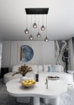 Lampa wisząca Cosmo 6-punktowa w kolorze grafitowym, z sześcioma przezroczystymi kloszami, zawieszona na czarnym stelażu. Ujęcie z góry, pokazujące lampę nad białym, owalnym stołem we wnętrzu.