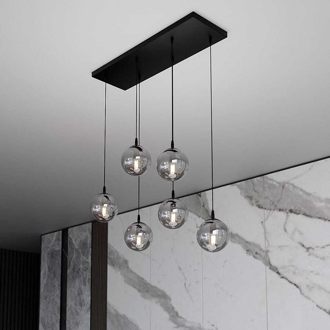 Lampa wisząca Cosmo 6-punktowa w kolorze grafitowym. Posiada sześć przezroczystych kulowych kloszy zawieszonych na czarnych przewodach, mocowanych do prostokątnej podstawy. Tło w odcieniach szarości z marmurowym wzorem.