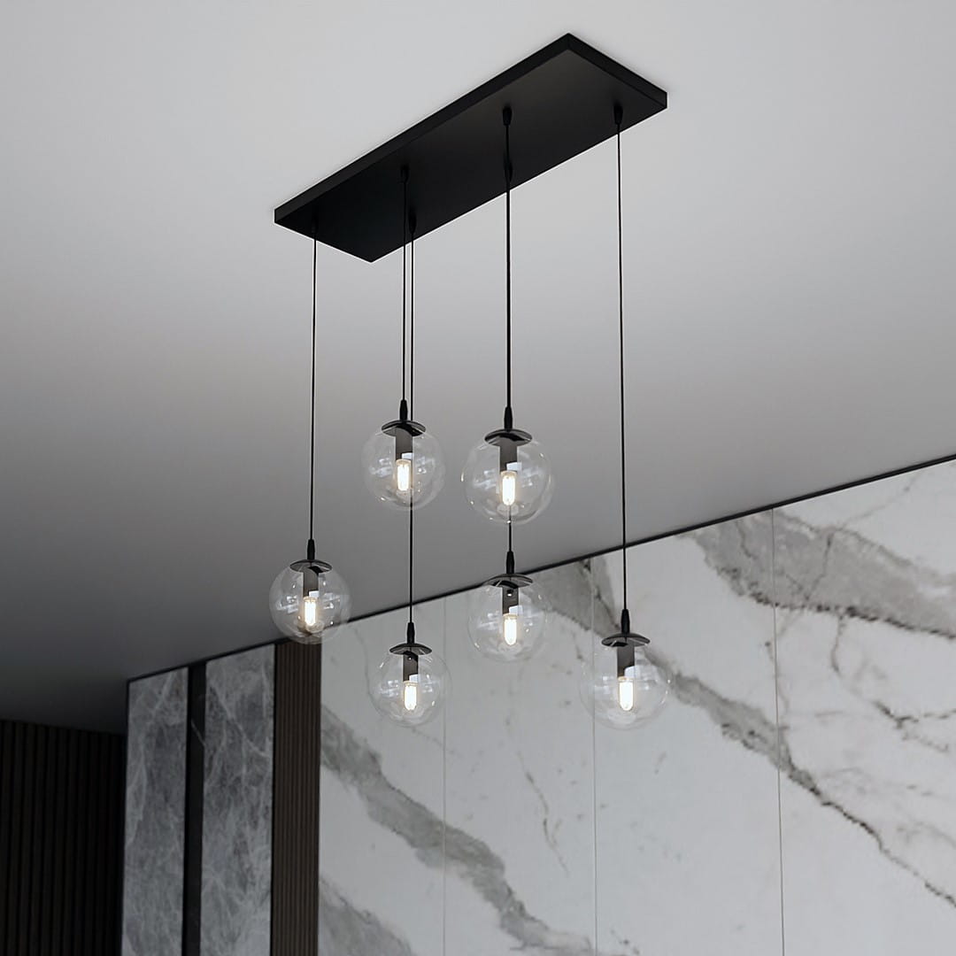 Lampa wisząca Cosmo 6-punktowa z sześcioma przezroczystymi kloszami w kształcie kul, zawieszona na cienkich czarnych kablach. Montaż na prostokątnej, czarnej podstawie. Tło w odcieniach szarości z marmurowym wzorem.