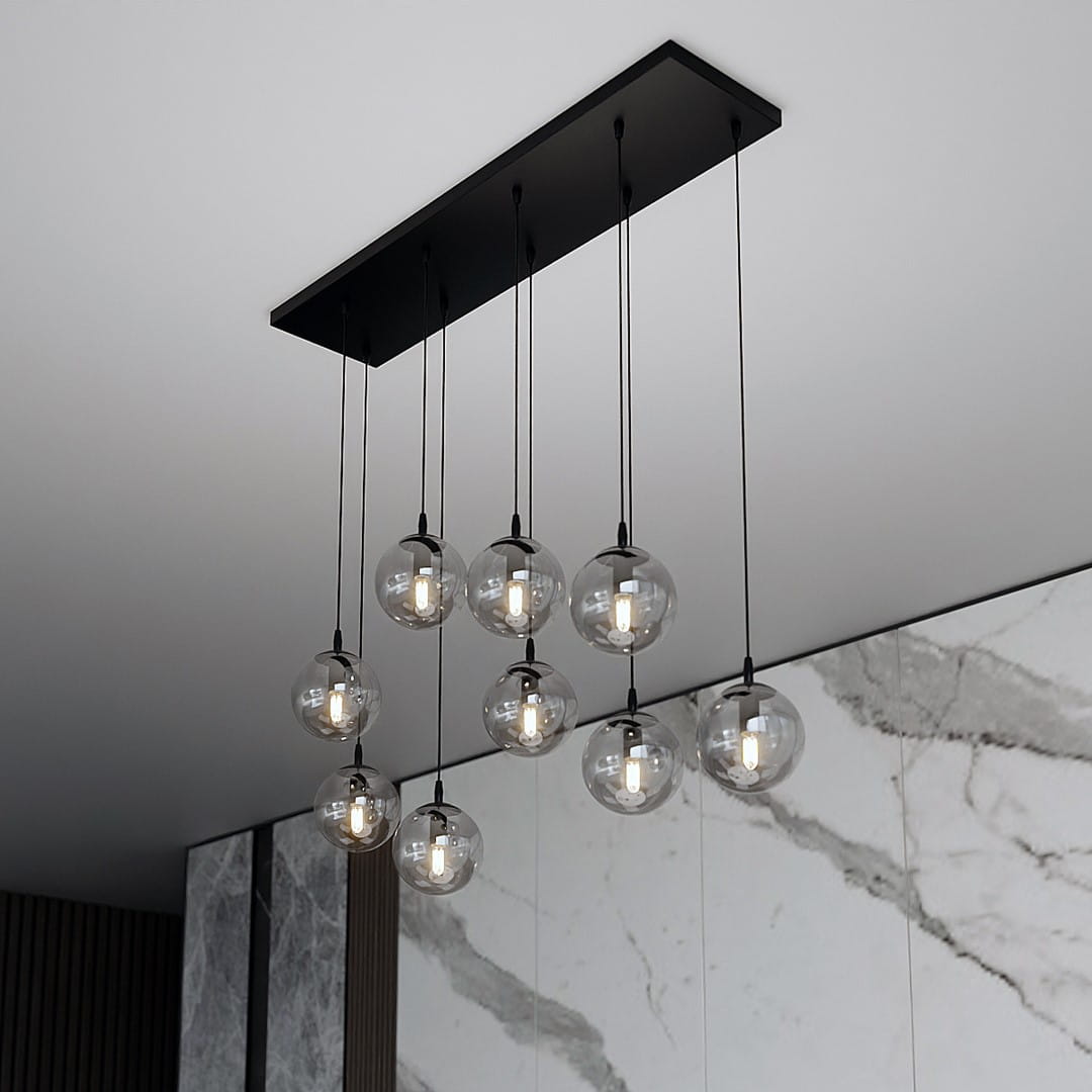 Lampa wisząca Cosmo 9-punktowa w kolorze grafitowym z ośmioma przezroczystymi kulkami szklanymi zawieszonymi na cienkich czarnych linkach, montowana na prostokątnej, czarnej podstawie. Tło stanowi jasna, marmurowa ściana.