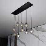 Lampa wisząca Cosmo 9-punktowa w kolorze grafitowym z ośmioma przezroczystymi kulkami szklanymi zawieszonymi na cienkich czarnych linkach, montowana na prostokątnej, czarnej podstawie. Tło stanowi jasna, marmurowa ściana.