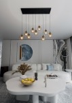 Lampa wisząca Cosmo 9-punktowa w kolorze miodowym, z transparentnymi kloszami, zawieszona na czarnym stelażu. Ujęcie z góry, pokazujące lampę w nowoczesnym wnętrzu z białym stołem oraz dekoracjami w tle.