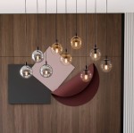 Lampa wisząca Cosmo Mix 1, 9-punktowa, z przezroczystymi i brązowymi kloszami w kształcie kul. Zawieszona na cienkich czarnych przewodach, w nowoczesnym wnętrzu z drewnianą ścianą i geometrycznymi dekoracjami w tle.