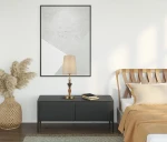 Lampa stołowa Moren w brązie antycznym z kloszem w kolorze beżowym, stojąca na ciemnym stoliku. W tle widoczny obraz w ramie oraz dekoracyjne elementy, jak kosz z roślinnością.