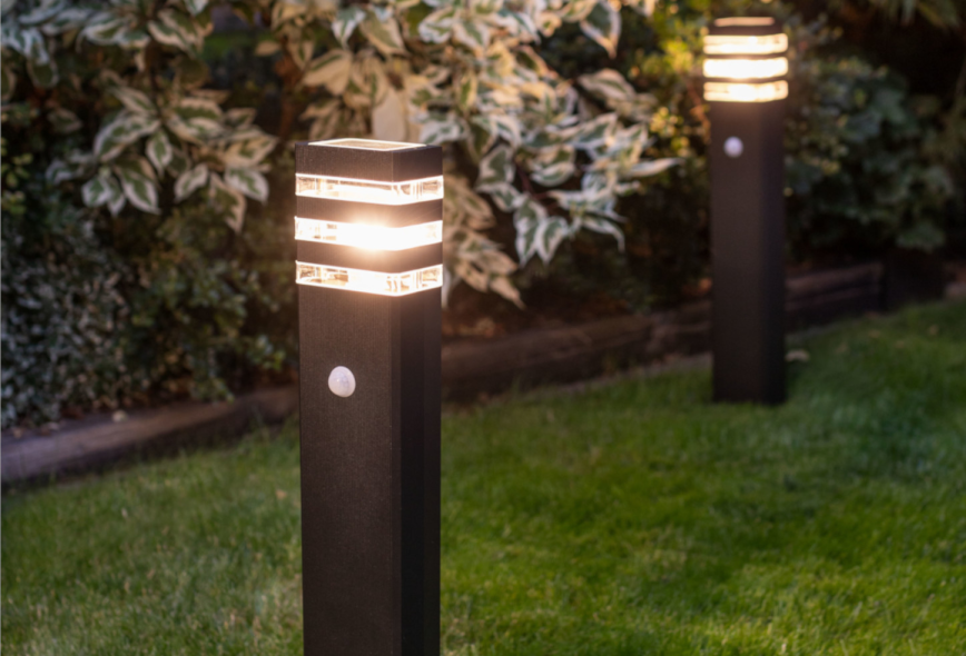 Lampa ogrodowa Rio Sensor, stojąca, z czujnikiem ruchu. Czarna, prostokątna obudowa z dekoracyjnymi, podświetlanymi paskami. Widoczny czujnik ruchu z boku. Umieszczona na trawniku, w tle roślinność.