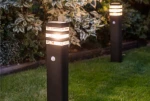 Lampa ogrodowa Rio Sensor, stojąca, z czujnikiem ruchu. Czarna, prostokątna obudowa z dekoracyjnymi, podświetlanymi paskami. Widoczny czujnik ruchu z boku. Umieszczona na trawniku, w tle roślinność.