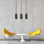 Lampa wisząca Tirana 3xE27 w kolorze złotym i czarnym, z trzema cylindrycznymi kloszami na smukłych przewodach, zawieszona w nowoczesnym wnętrzu z żółtymi krzesłami i białym stołem. Tło w odcieniu szarości.