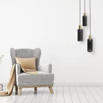 Lampa wisząca Tirana 3xE27 w kolorze złotym i czarnym, z trzema cylindrycznymi kloszami o siatkowanej fakturze, zawieszona nad szarym fotelem z beżową poduszką, na tle białej ściany. Kable w czarnym kolorze, minimalistyczny design.