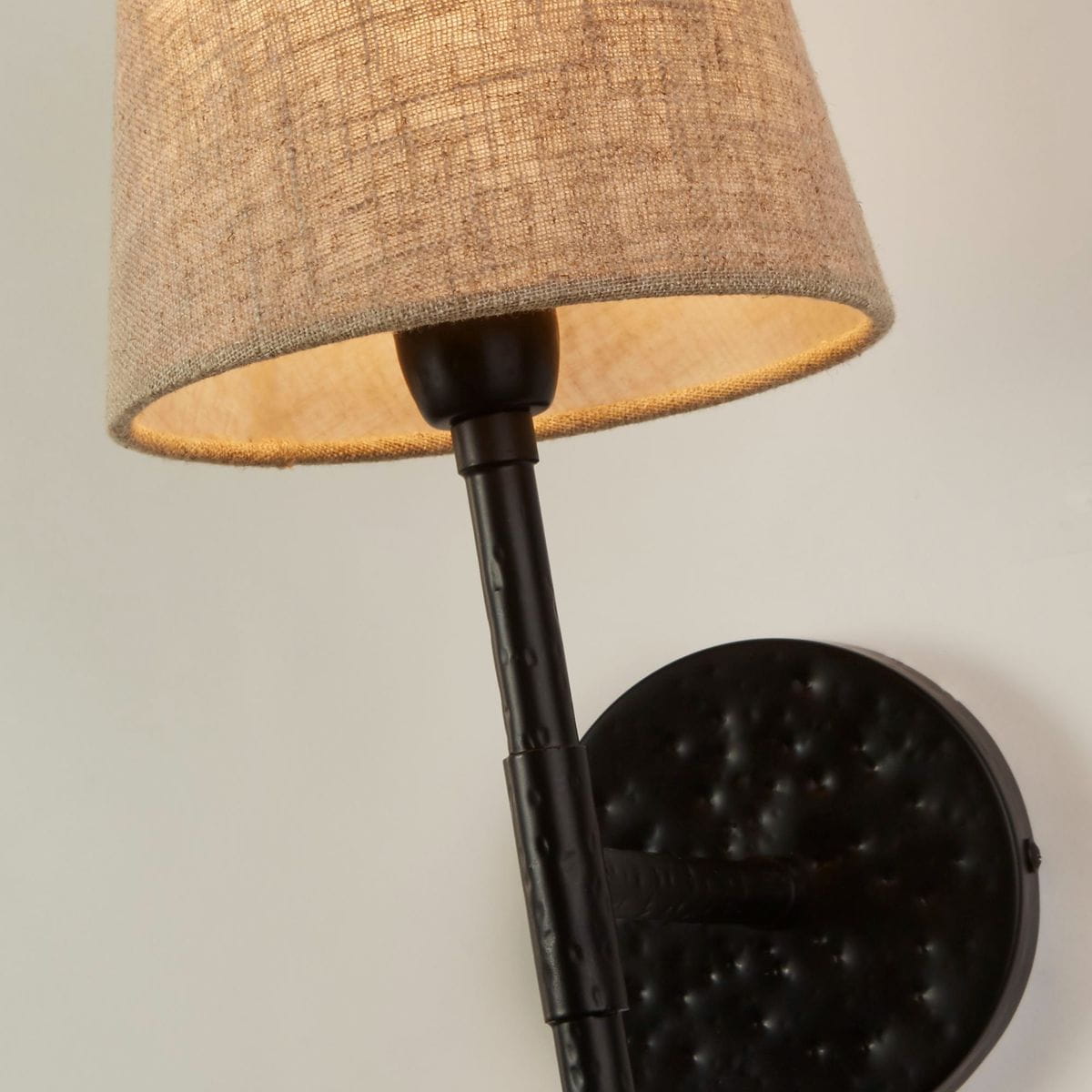 Kinkiet Gothic w czarnym kolorze z abażurem z materiału w odcieniu naturalnego lnu. Lampa zamontowana na ścianie, z prostym, metalowym ramieniem i okrągłą podstawą. Widoczne detale teksturowane na metalowej części.