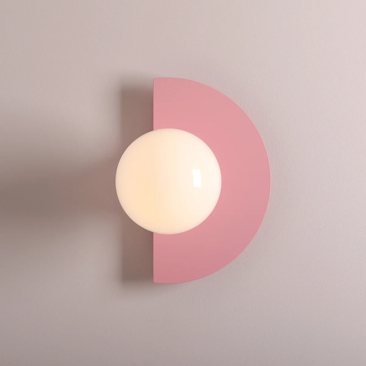 Kinkiet Loop Baby Pink z okrągłą, mlecznobiałą kulą umieszczoną na różowym, półokrągłym tle. Zamontowany na gładkiej, jasnoszarej ścianie, podkreślający nowoczesny design i subtelną kolorystykę. Idealny do wnętrz dziecięcych lub stylowych aranżacji.