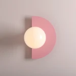 Kinkiet Loop Baby Pink z okrągłą, mlecznobiałą kulą umieszczoną na różowym, półokrągłym tle. Zamontowany na gładkiej, jasnoszarej ścianie, podkreślający nowoczesny design i subtelną kolorystykę. Idealny do wnętrz dziecięcych lub stylowych aranżacji.