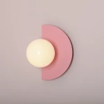 Kinkiet Loop Baby Pink z jednym źródłem światła E14, o okrągłej, matowej, białej kulistej kloszu, umieszczony na różowym tle w formie półkola. Produkt montowany na ścianie, widok frontalny.