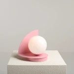 Lampa stołowa Loop Baby Pink z okrągłą, białą żarówką, na różowej podstawie z geometrycznym, półkolistym ekranem. Ujęcie frontalne, produkt stoi na teksturowanej powierzchni.