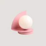 Lampa stołowa Loop Baby Pink z okrągłą, białą kulą na różowej podstawie i półokrągłym ekranem. Ujęcie z boku, ukazuje nowoczesny design. Idealna do aranżacji wnętrz.