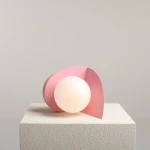 Lampa stołowa Loop w kolorze baby pink z okrągłym, mlecznym kloszem. Ułożona na białym, teksturowanym podłożu, z subtelnym tłem. Kształt lampy jest nowoczesny, z zaokrąglonymi krawędziami i dynamiczną formą.
