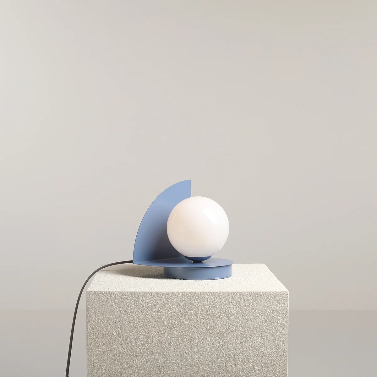 Lampa stołowa Loop Dusty Blue z okrągłą, białą żarówką na niebieskiej podstawie w kształcie półokręgu. Ujęcie z boku, produkt stoi na białym, teksturowanym podłożu. Widoczny kabel zasilający. Tło jednolite, jasne.
