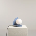 Lampa stołowa Loop Dusty Blue z okrągłą, białą żarówką na niebieskiej podstawie w kształcie półokręgu. Ujęcie z boku, produkt stoi na białym, teksturowanym podłożu. Widoczny kabel zasilający. Tło jednolite, jasne.