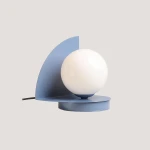 Lampa stołowa Loop w kolorze błękitnym, z okrągłą, białą żarówką. Ma nowoczesny kształt z asymetrycznym, półokrągłym panelu oraz okrągłą podstawą. Widoczny czarny kabel. Idealna do minimalistycznych wnętrz.