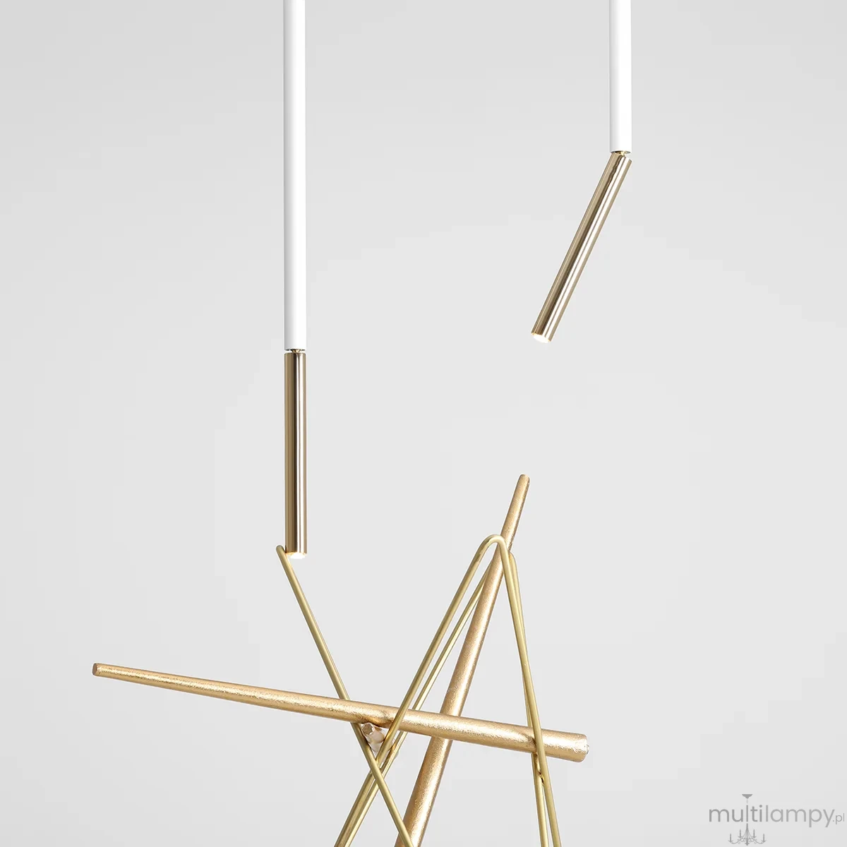 Lampa sufitowa Stick S w białym i złotym kolorze, z metalowymi elementami. Kompozycja składa się z białych rur i złotych prętów ułożonych w geometryczny kształt. Wisi w pionie na jednolitym, jasnym tle.