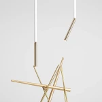 Lampa sufitowa Stick S w białym i złotym kolorze, z metalowymi elementami. Kompozycja składa się z białych rur i złotych prętów ułożonych w geometryczny kształt. Wisi w pionie na jednolitym, jasnym tle.